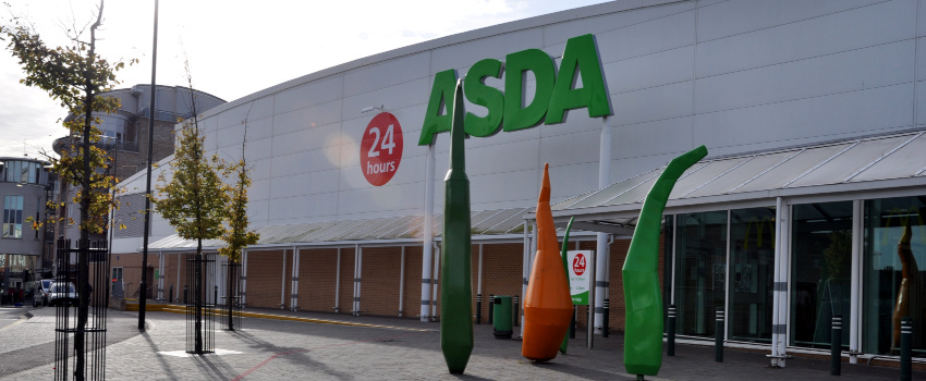 A long modern building with the word Asda in large green letters; in front of the building are small trees and three large model vegetables