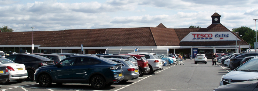 A wide, short building with brown roof tiles and the words 'Tesco Extra' in large lettering, and a car park in front.